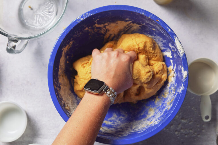Someone with a watch kneads dough for pumpkin cinnamon rolls in a blue bowl, with cups and a glass bowl nearby on a light surface.