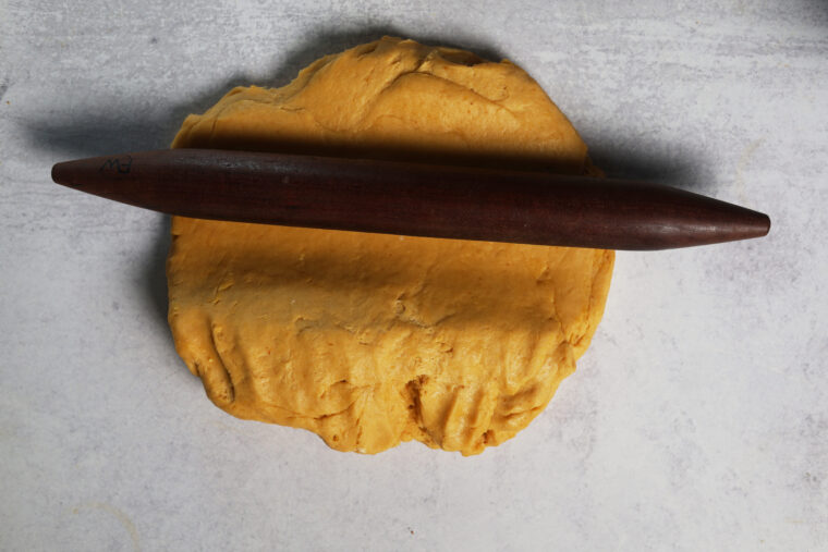 A dark wooden rolling pin sits at an angle on some yellow pumpkin roll dough, all on a light gray counter.