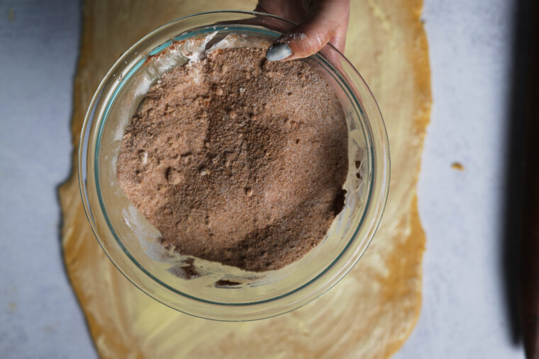 Someone’s holding a bowl of cinnamon sugar over buttery dough, all set to fill up for some tasty pumpkin cinnamon rolls.