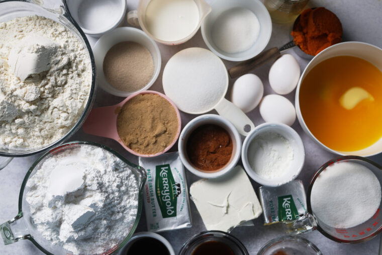 Overhead shot of bowls and cups filled with flour, sugars, eggs, butter, cream cheese, pumpkin puree, milk, vanilla & baking powder.